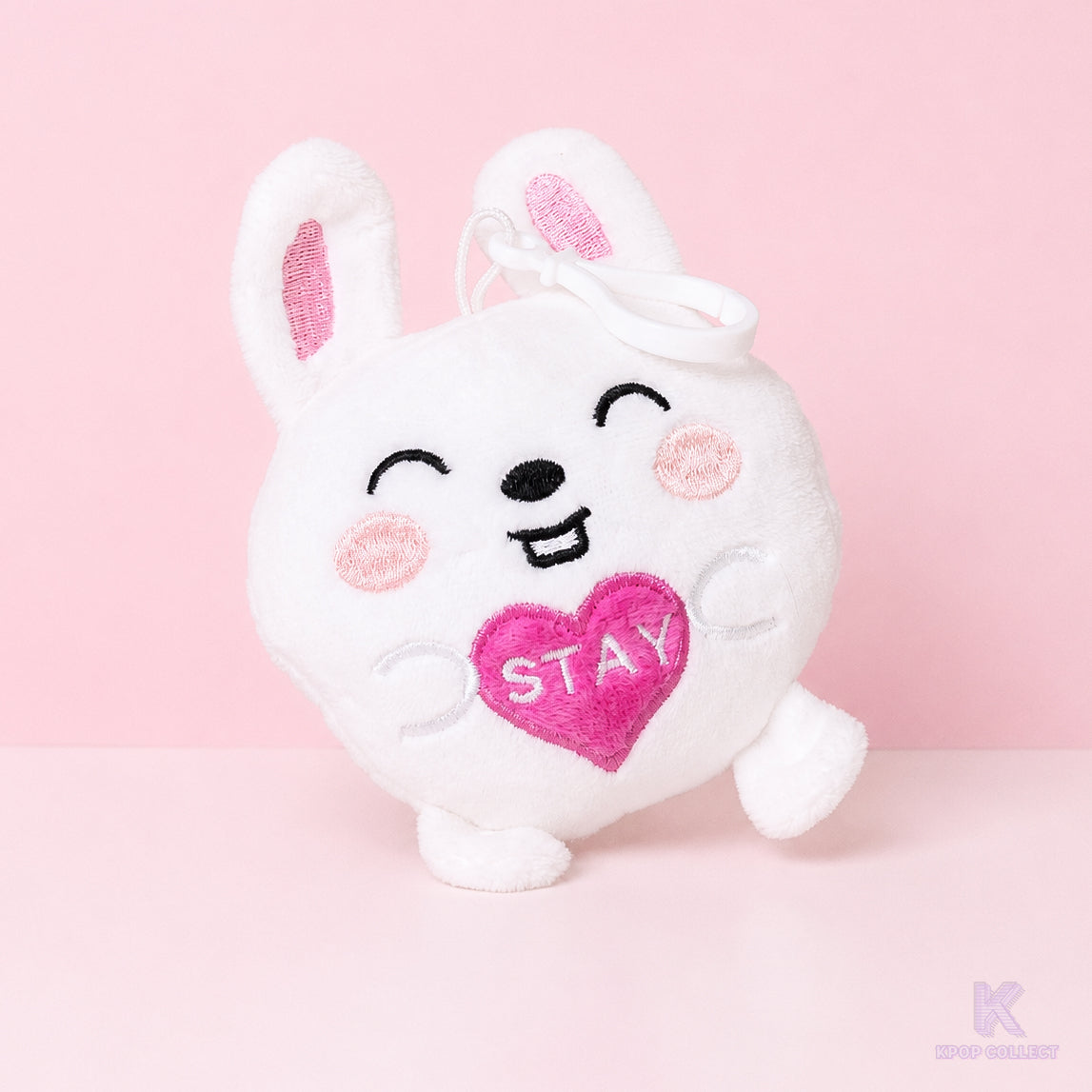 A direct, front-facing view of the Stray Kids Lee Know Leebit plush keychain. The white bunny character is holding a small pink heart with the word STAY on it against a soft pink background.
