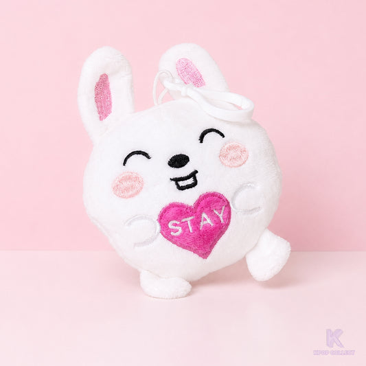 A direct, front-facing view of the Stray Kids Lee Know Leebit plush keychain. The white bunny character is holding a small pink heart with the word STAY on it against a soft pink background.